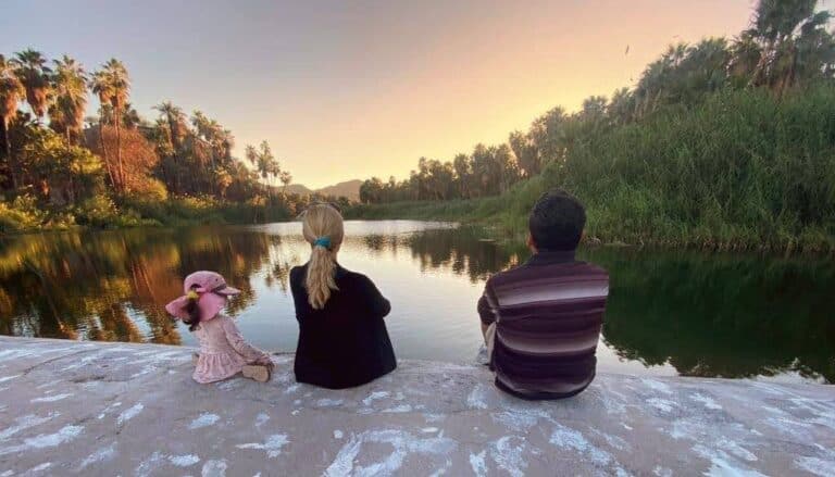 Three people, two adults and a little girl, enjoy a view of a lake at sunset with their backs to the camera.