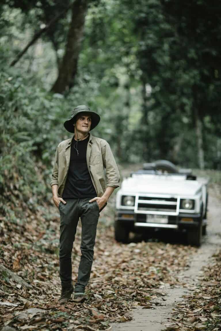 Positive young man walking in autumnal forest near car parked on narrow path